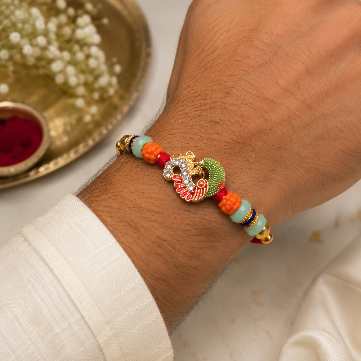 A man's arm wears a red thread with a gold and green peacock charm, orange and mint green beads, and gold accents, with a