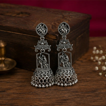A pair of oxidized silver jhumka earrings with Radha Krishna temple design on a wooden surface.