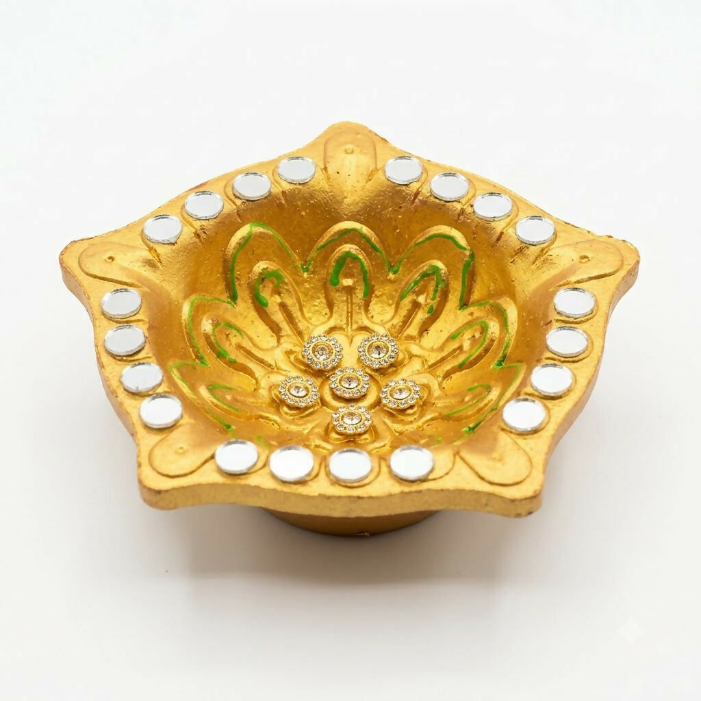 A close-up of a gold star-shaped diya with green painted details, small mirrors, and rhinestone flowers.