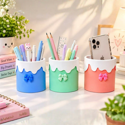 Three pastel organizers on a desk, filled with writing instruments and a phone, beside a pink notebook.