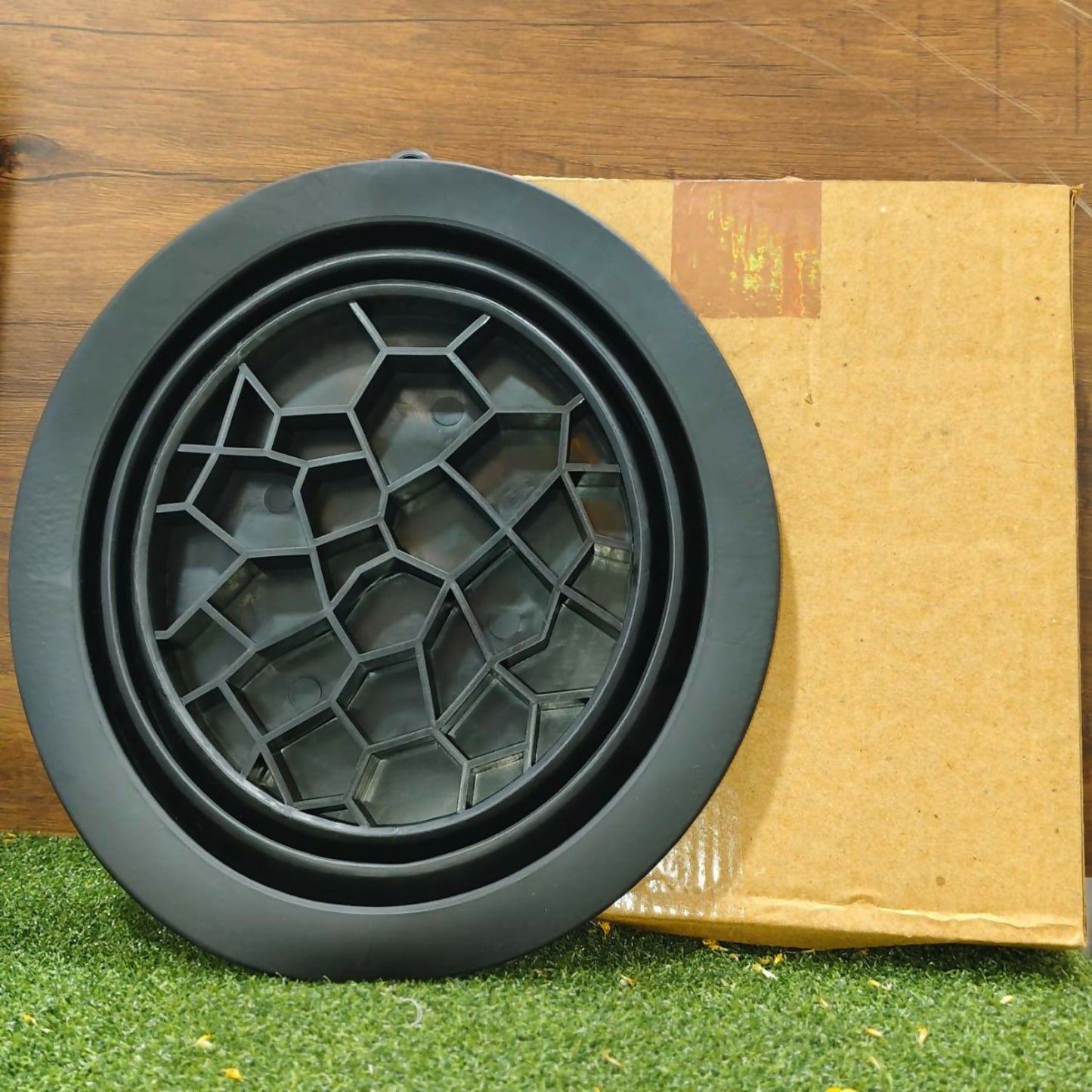 A black silicone ice mold with a geometric pattern on the lid rests on artificial grass next to a cardboard box.