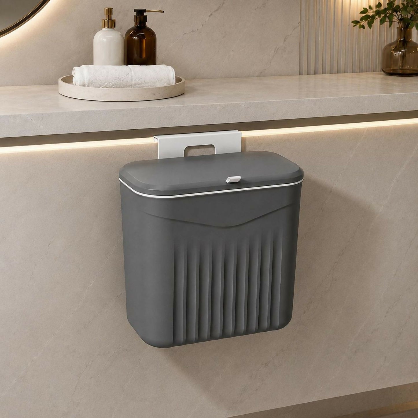A dark grey plastic dustbin with a lid hangs from a white hook on a light grey cabinet in a bathroom setting.