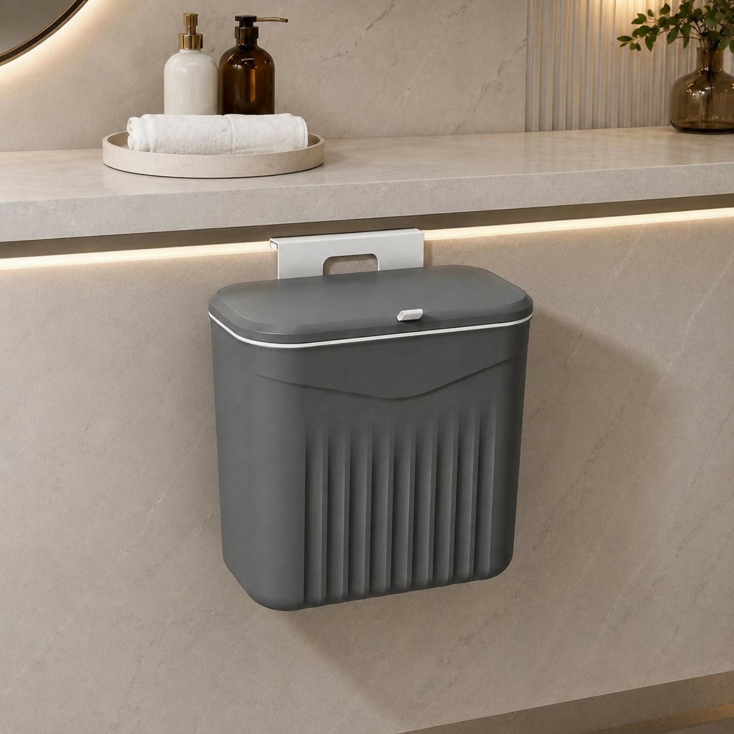 A dark grey plastic dustbin with a lid hangs from a white hook on a light grey cabinet in a bathroom setting.