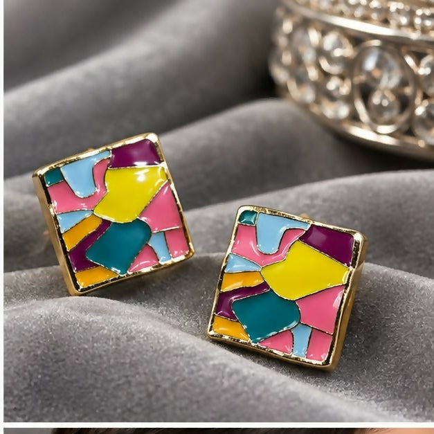 A pair of square stud earrings with abstract multicolour enamel and gold borders are angled on grey fabric.