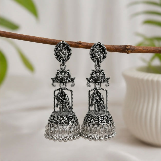 A pair of oxidized silver jhumka earrings with Radha Krishna temple design hanging from a tree branch.