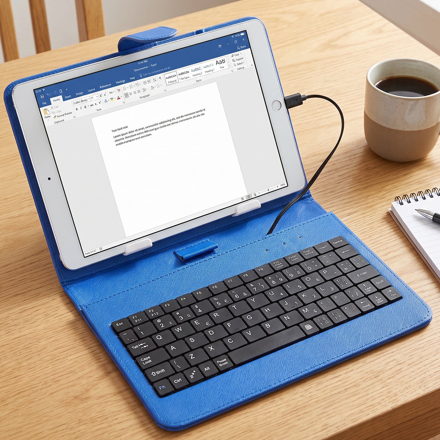 A blue PU leather case with a tablet and wired keyboard sits on a wooden table next to a coffee mug.