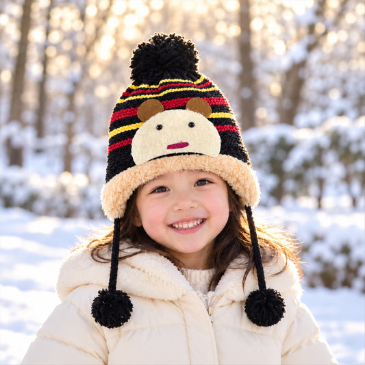 A smiling child wears a black, red, and yellow striped knitted hat with a cream animal face and brown ears, in a snowy