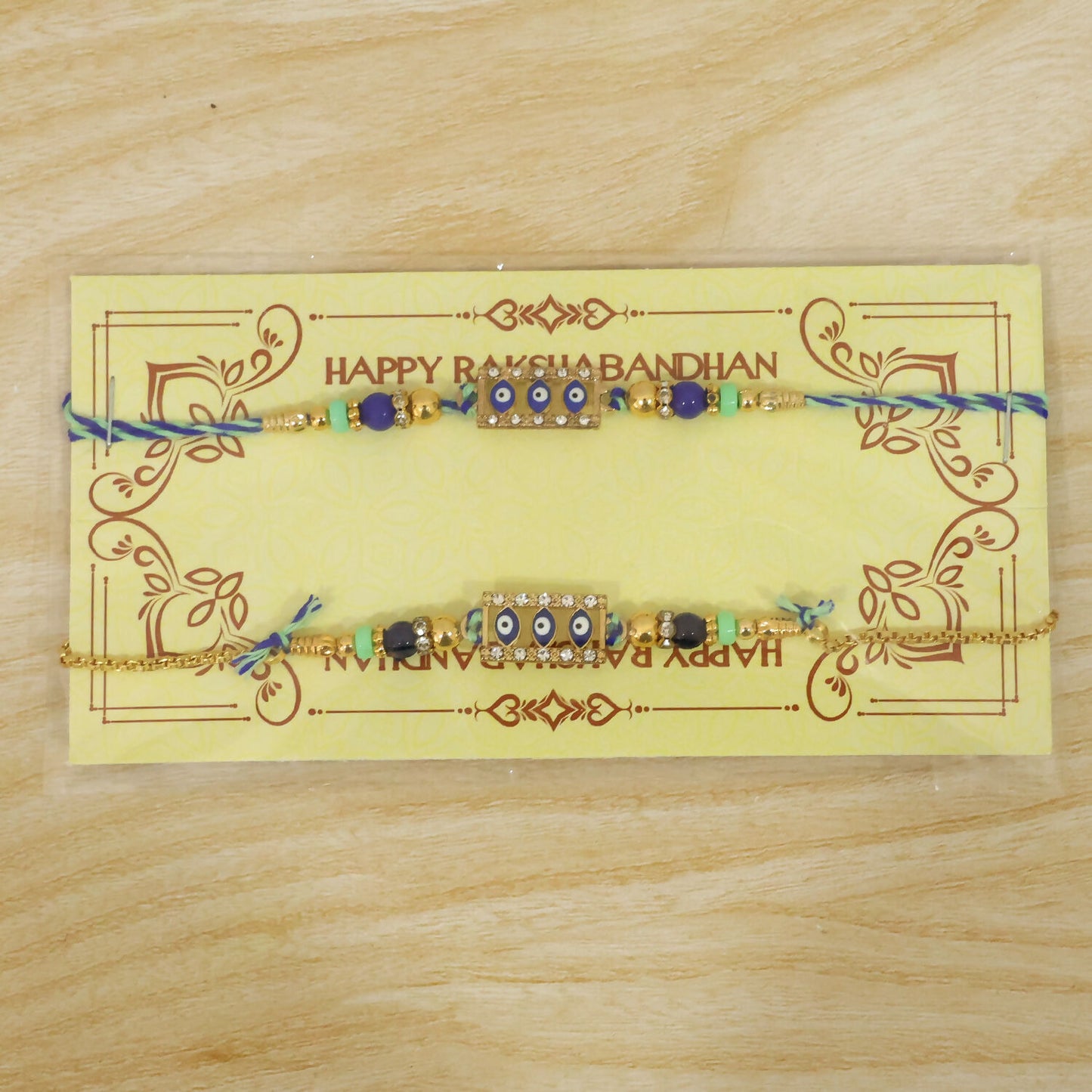 Two blue and gold evil eye rakhis, one a bracelet, on a yellow Happy Rakshabandhan card.