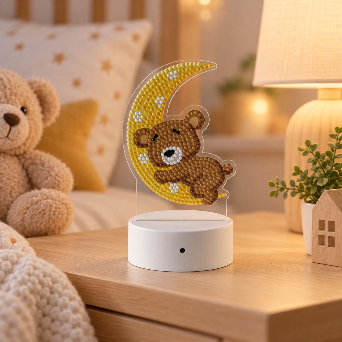 The teddy bear and moon diamond painting night light on a wooden table, next to a large teddy bear and a white lamp.