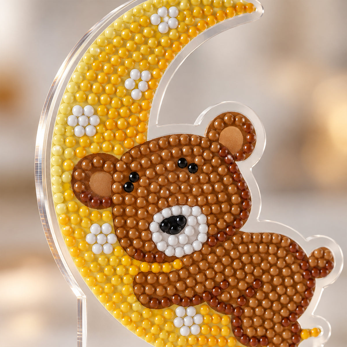 A close-up of the diamond painting, showing the brown teddy bear with black eyes and nose, resting on a yellow moon with