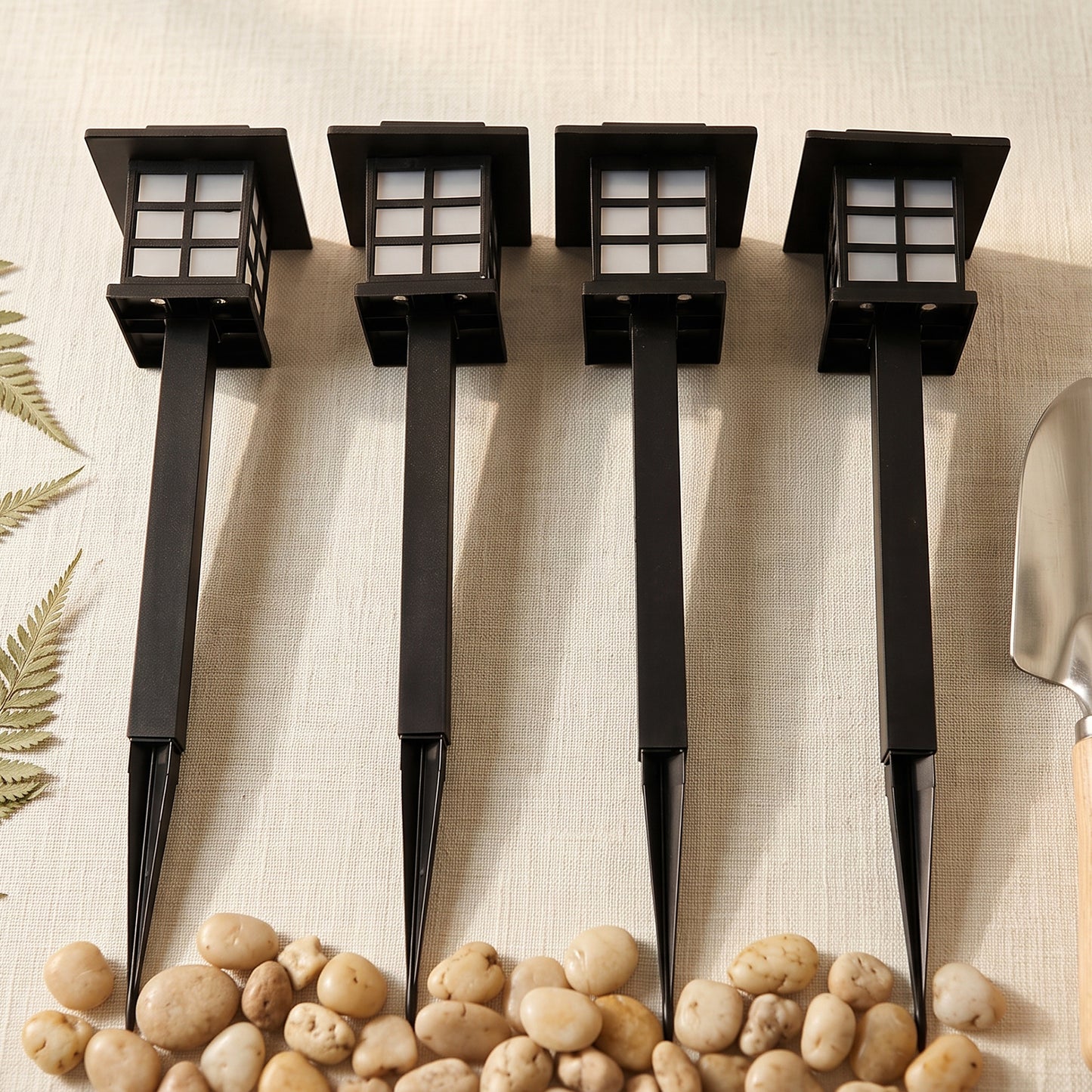 Four black solar garden lights with stakes are lined up on a light surface with pebbles and a small shovel.