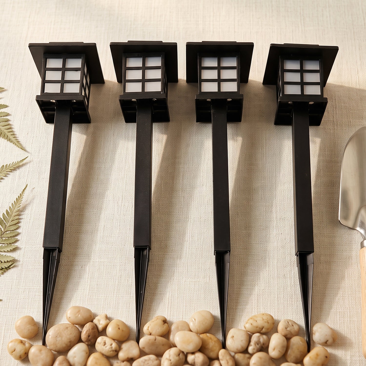 Four black solar garden lights with stakes are lined up on a light surface with pebbles and a small shovel.