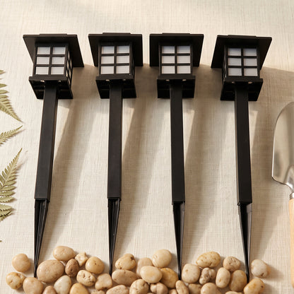 Four black solar garden lights with stakes are lined up on a light surface with pebbles and a small shovel.