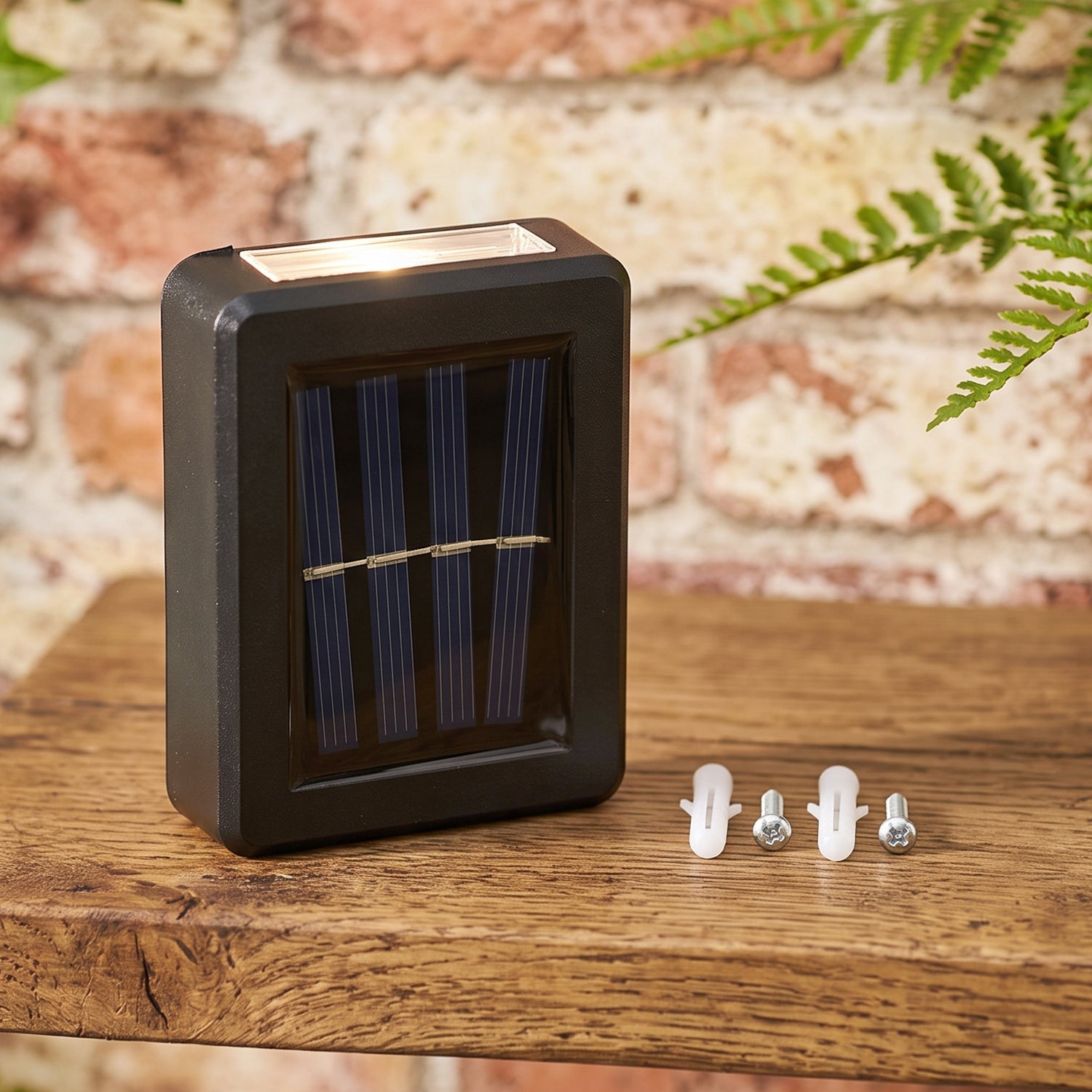 A black solar LED wall light with a blue solar panel sits on a wooden surface next to two white wall plugs and two silver