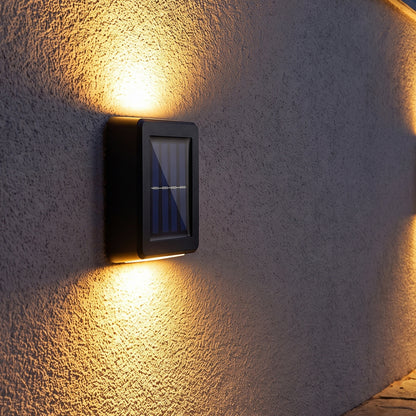 Two black solar LED wall lights illuminate a grey textured wall with warm light beams pointing up and down in an outdoor