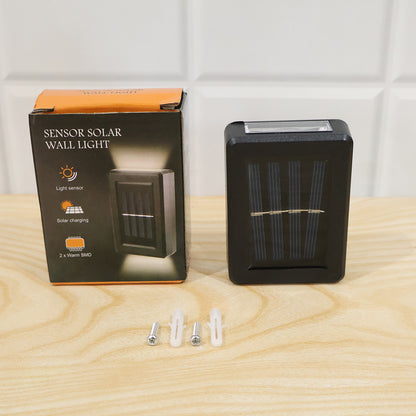 A black solar LED wall light and its product box with a light sensor icon are displayed on a wooden surface with mounting