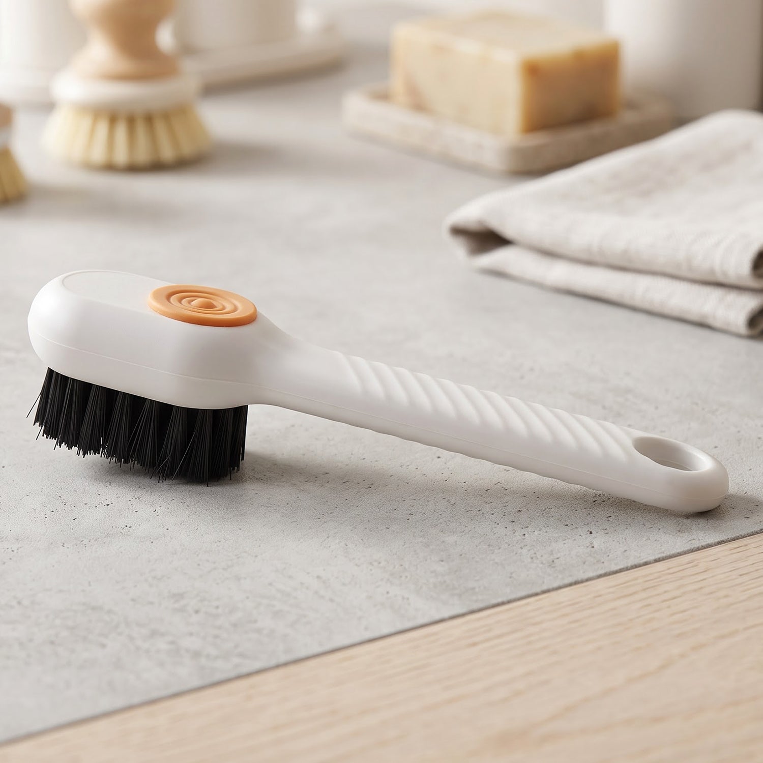 A white scrubbing brush with black bristles and an orange button rests on a gray surface with other cleaning items.