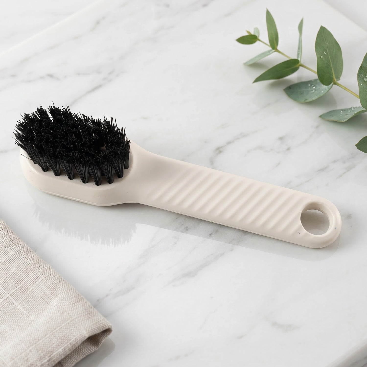 A beige scrubbing brush with black bristles rests on a white marble surface next to a folded linen cloth and green leaves.