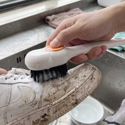 A hand holds a white scrubbing brush with an orange button, cleaning a mud-splattered white sneaker over a sink.