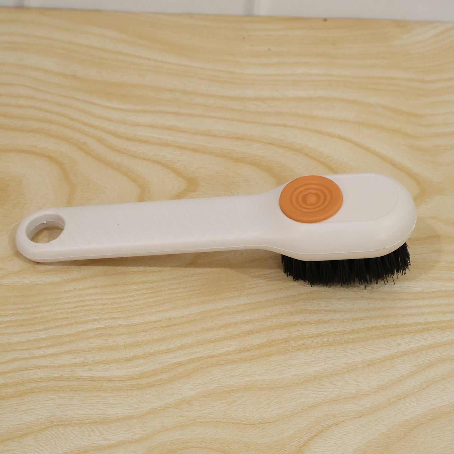 A white scrubbing brush with black bristles and an orange button lies on a light wooden surface.