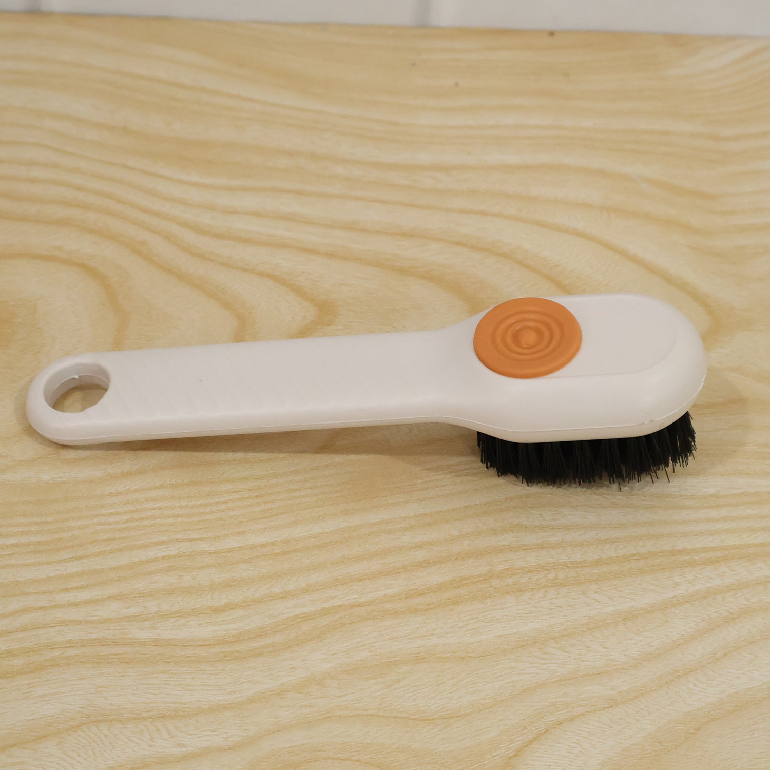 A white scrubbing brush with black bristles and an orange button lies on a light wooden surface.