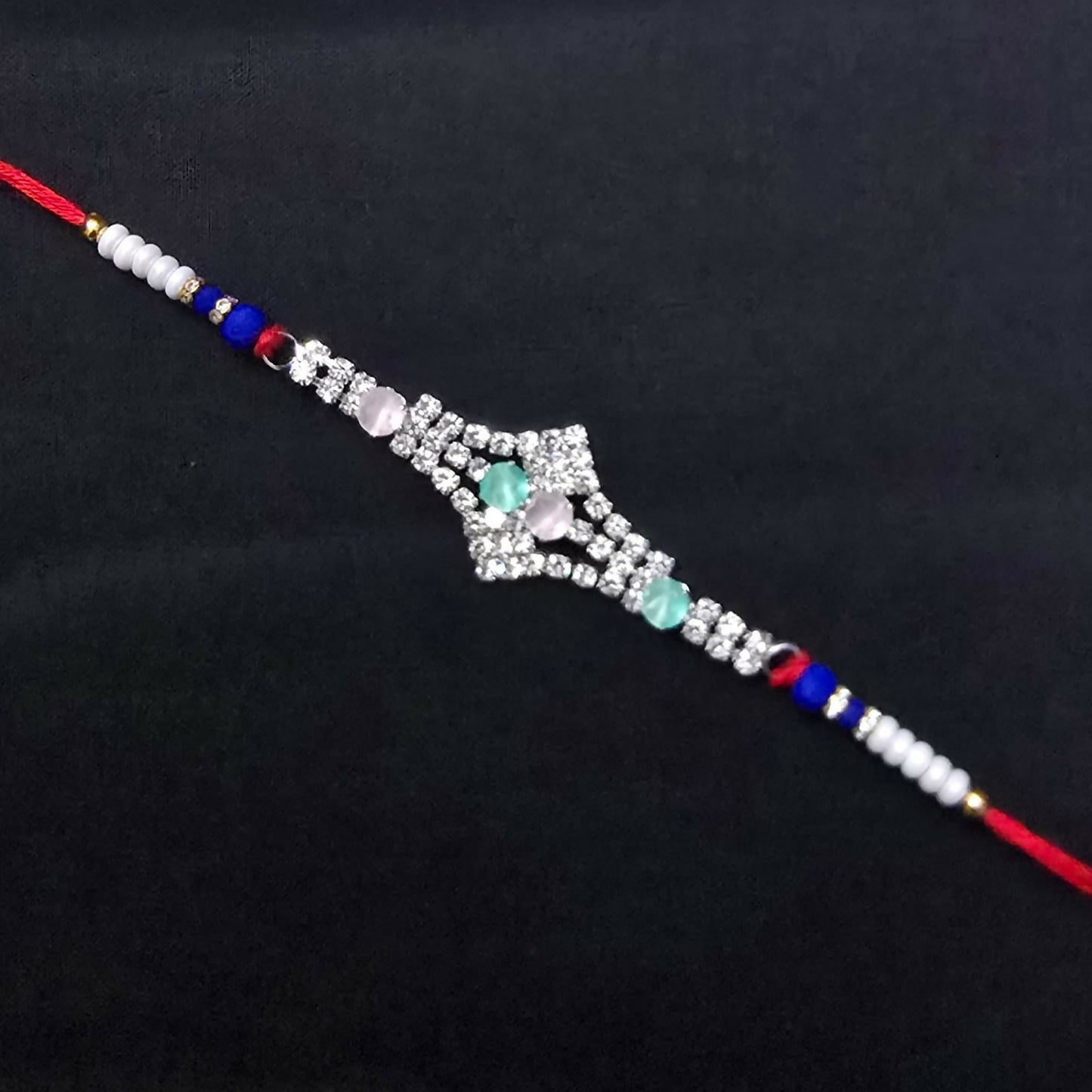 A red thread bracelet with a silver-toned, crystal-studded centerpiece and pink and green oval stones is displayed on a