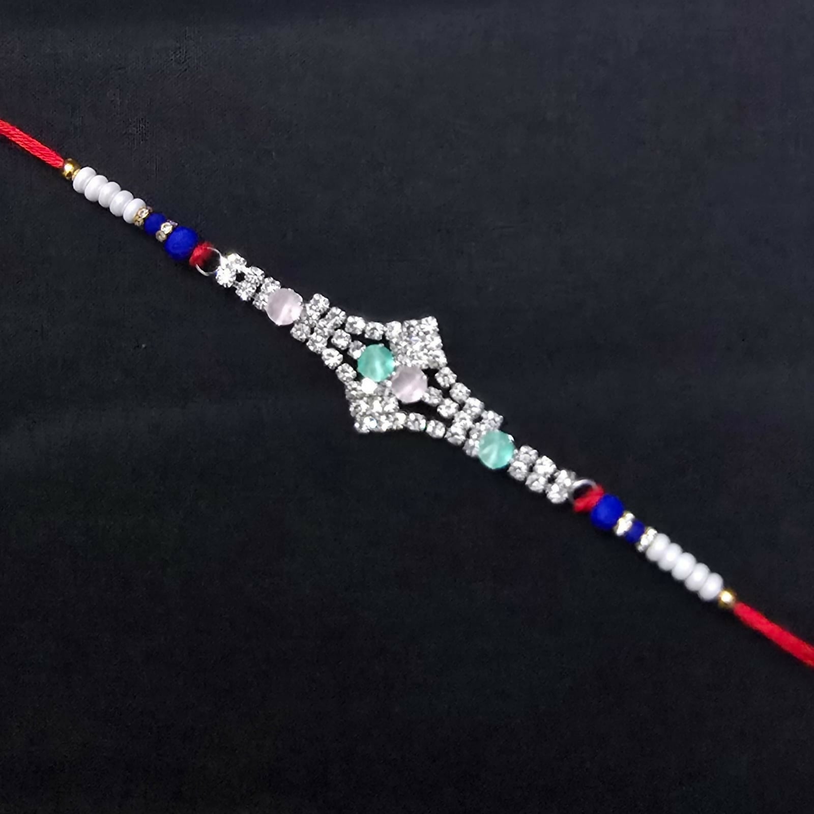 A red thread bracelet with a silver-toned, crystal-studded centerpiece and pink and green oval stones is displayed on a