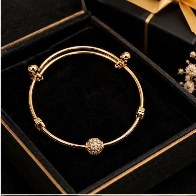 A gold-tone adjustable bangle with a crystal ball charm rests inside a black gift box with a gold ribbon.