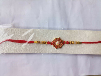 A red and gold beaded bracelet with a central red floral medallion and white pearl, displayed on an embossed white card