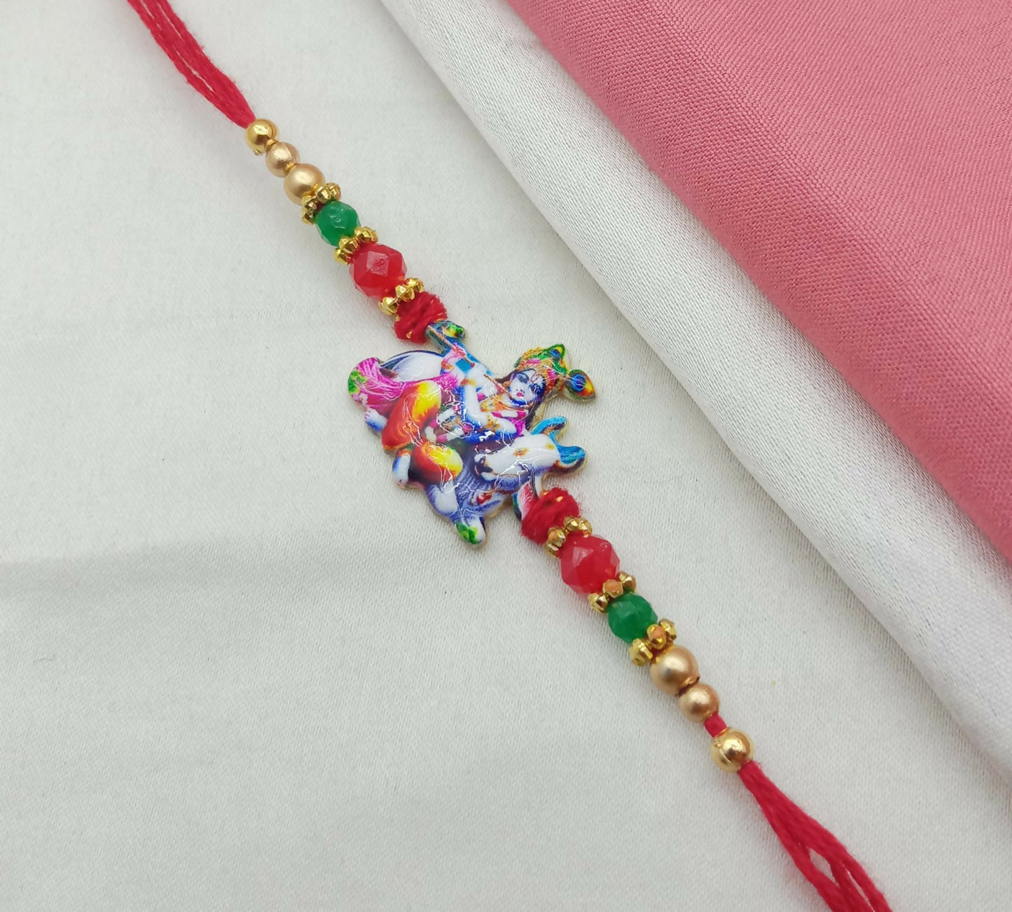 A red thread with a colorful Bal Gopal Krishna and cow charm, flanked by red, green, and gold beads, is laid diagonally