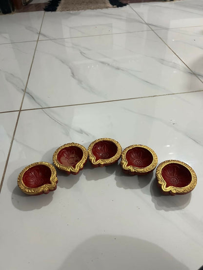 Five red and gold clay diyas with embossed patterns are neatly arranged in a row on a white tiled floor.