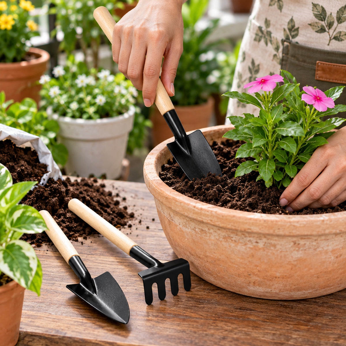 A person uses a small black trowel with a wooden handle to plant a pink flower in a terracotta pot.