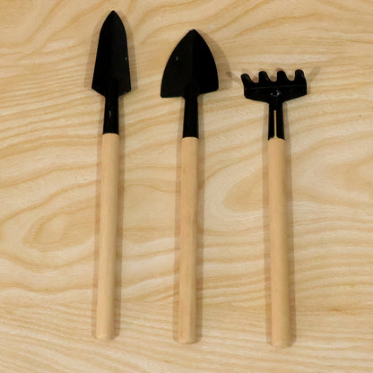 Two small black trowels and a matching rake with light wooden handles are neatly arranged on a wooden surface.