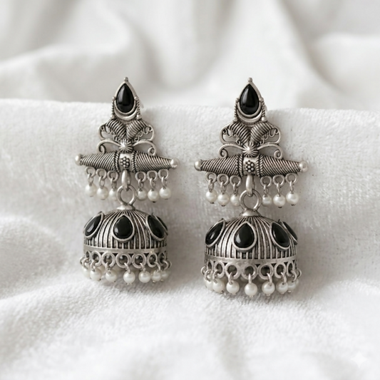 A pair of ornate oxidized silver jhumka earrings with black teardrop stones and white pearl accents on a white textured