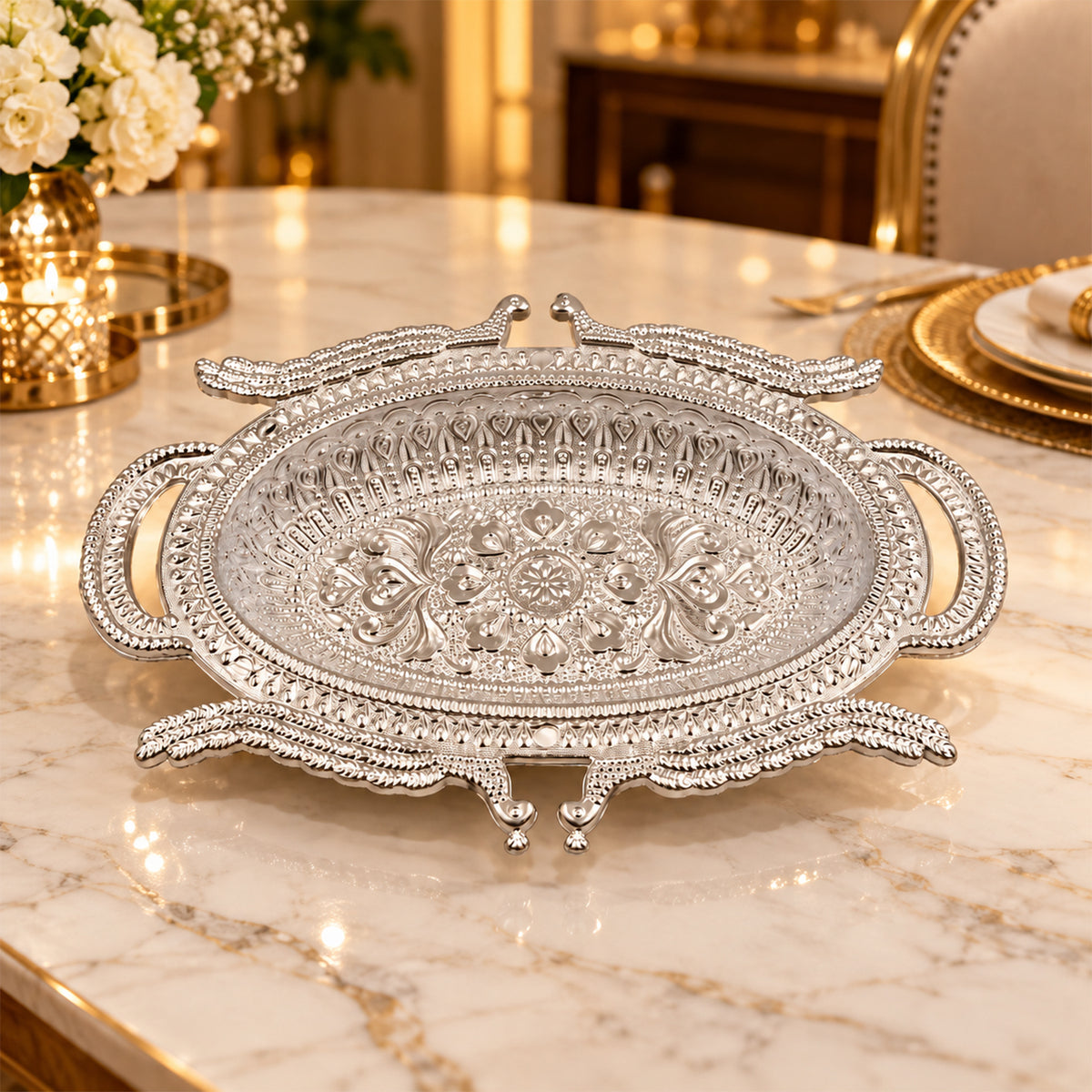 Empty silver-colored ornate serving tray on a marble table with blurred golden decor and flowers in the background.