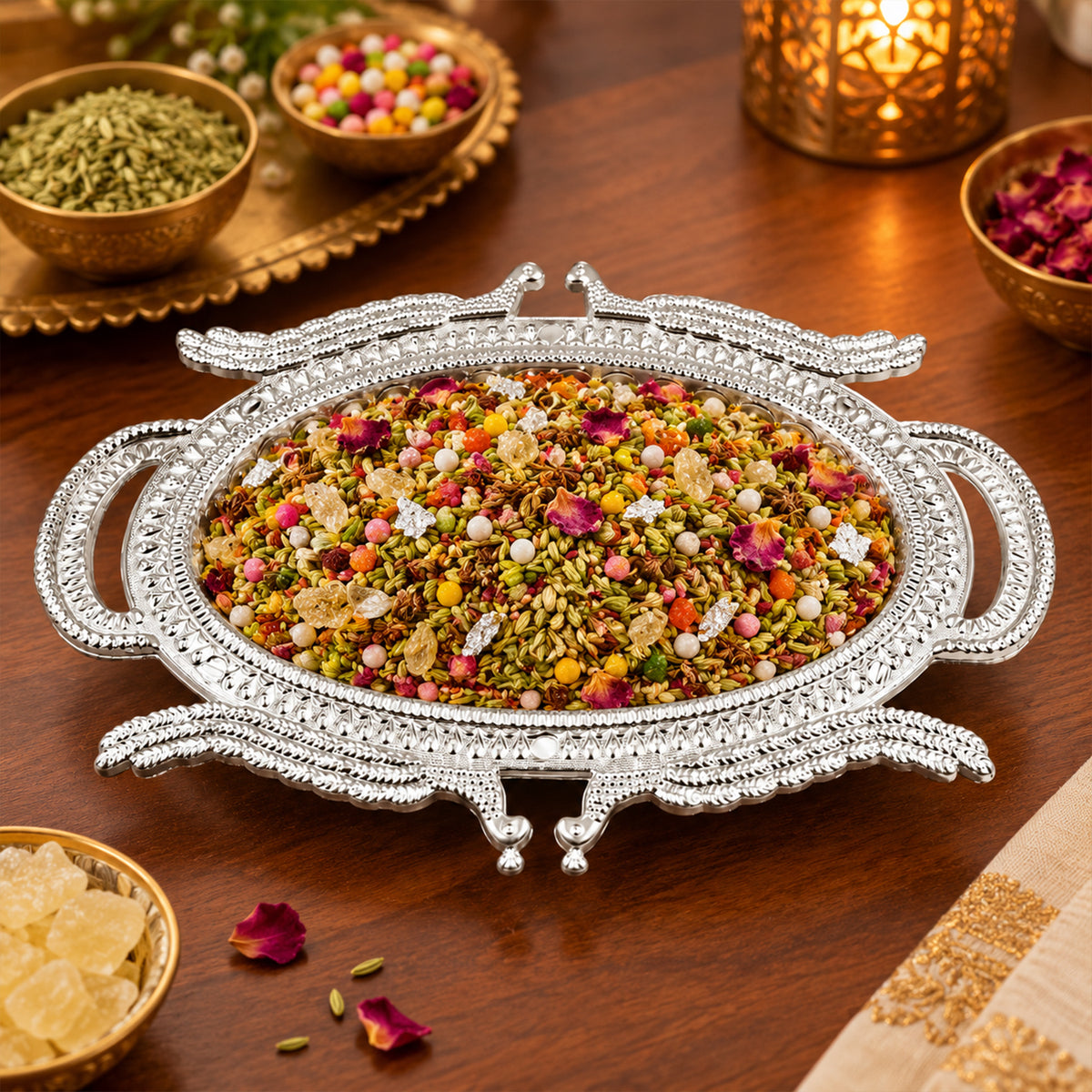 Ornate silver-colored serving tray filled with mukhwas, surrounded by small bowls and scattered petals on a table.