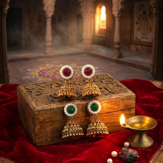 Two pairs of antique gold jhumka earrings with red and green stones, surrounded by pearls, on a wooden box.