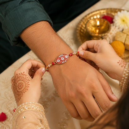 A person's hand ties a red and gold Rakhi onto another person's wrist, with a lit lamp and sweets in the background.
