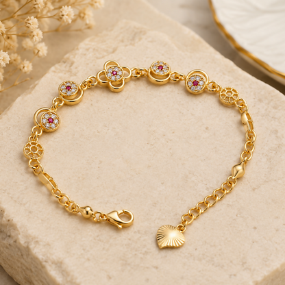 A gold-plated bracelet with floral-shaped charms, red and clear stones, lies on a light beige textured surface.