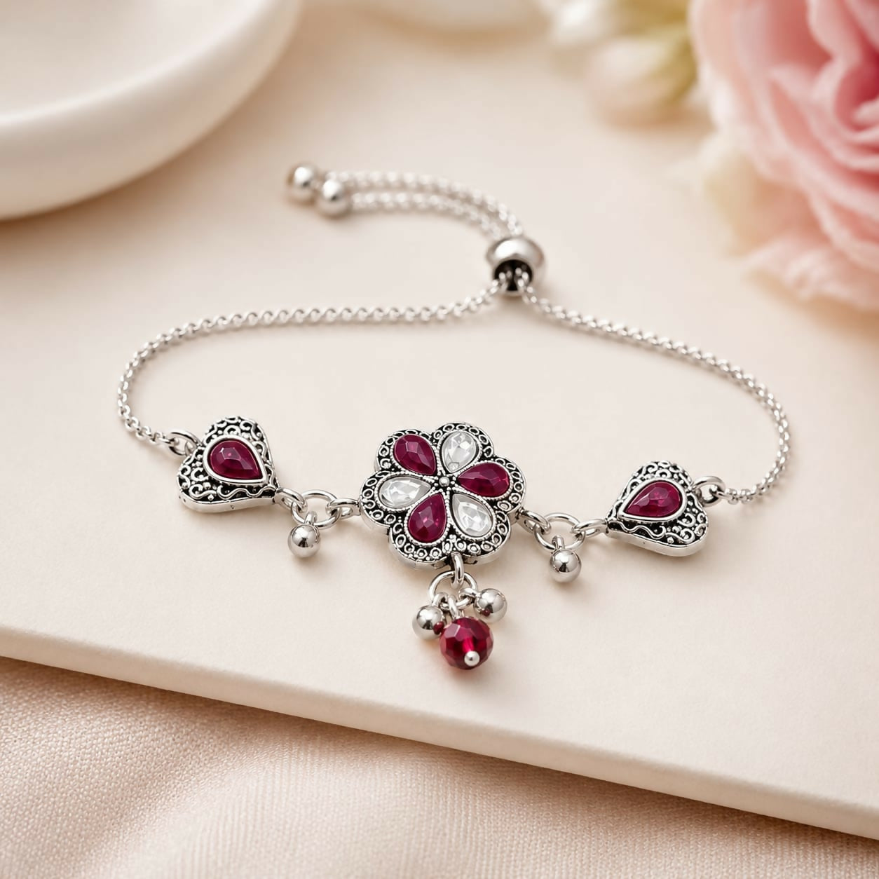 An antique silver adjustable slider bracelet with a central flower, two hearts, ruby-red pear stones, and dangling beads