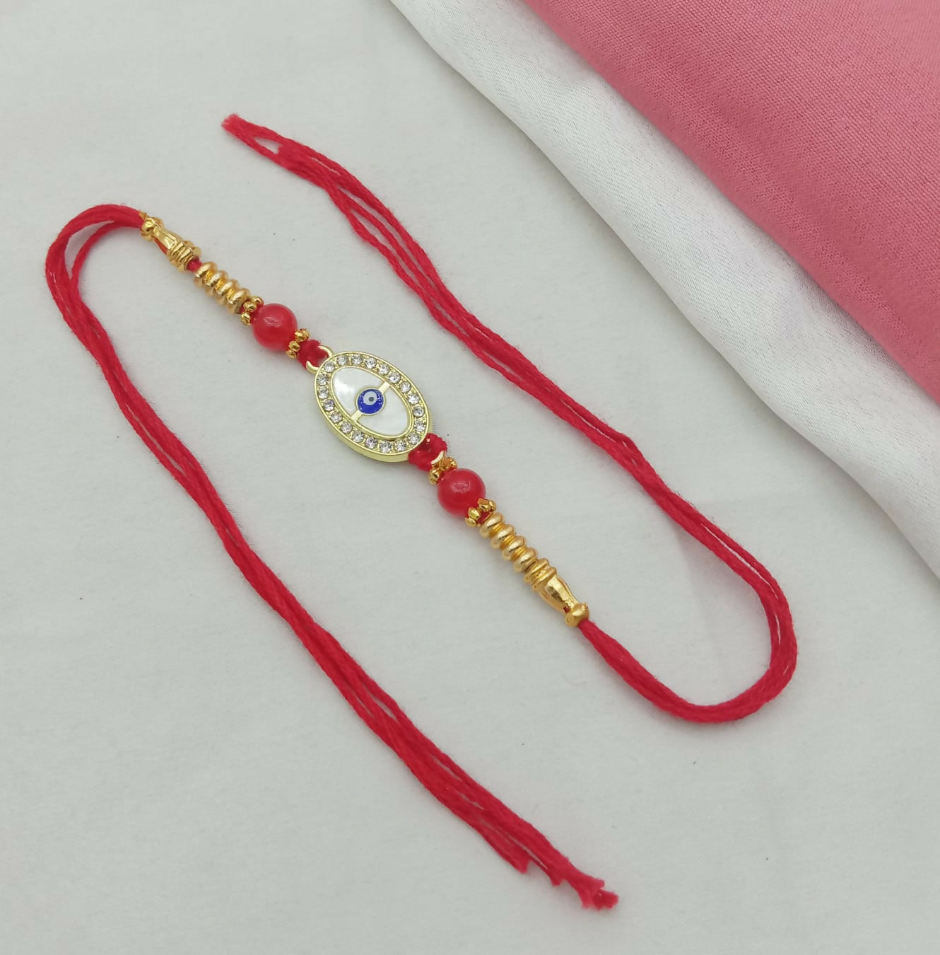 A red thread bracelet with a golden oval evil eye charm, white stones, and red beads lies on a white surface with a pink and