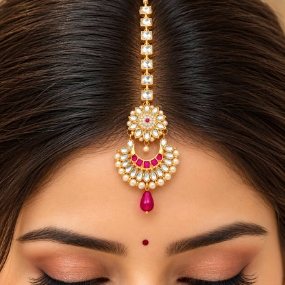 A close-up of a woman's forehead shows a gold maang tikka with clear and pink gems, pearls, and a single fuchsia drop.