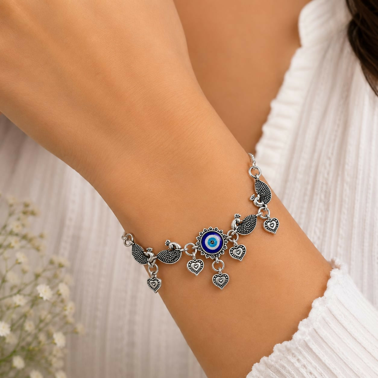 A woman wears the oxidized silver evil eye peacock charm bracelet with heart dangles on her wrist, against a white shirt.