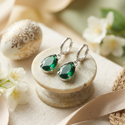 Green teardrop crystal earrings with silver hooks displayed on a light marble stand, surrounded by flowers and ribbon.