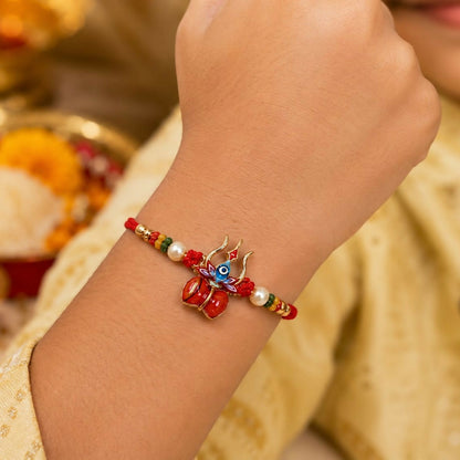 A child's arm wears a red thread bracelet featuring a gold trishul and damru charm with blue enamel, flanked by pearls and