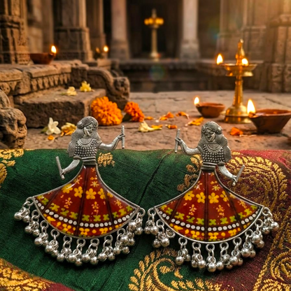 A pair of silver oxidized dancing girl earrings with maroon and yellow skirts, displayed on green fabric with lit diyas in