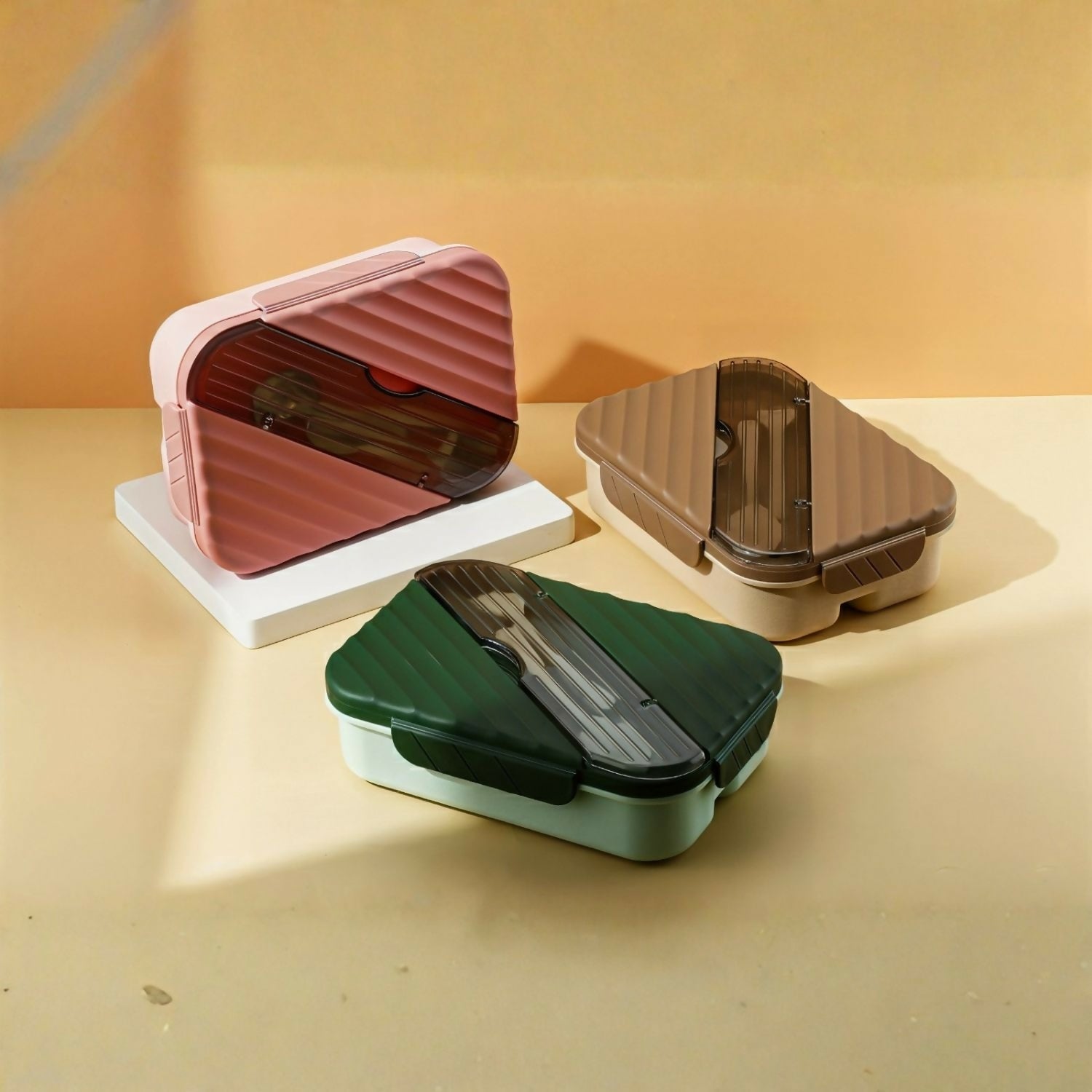 Three closed bento lunch boxes in pink, brown, and green with clear lids revealing cutlery, sitting on white and beige