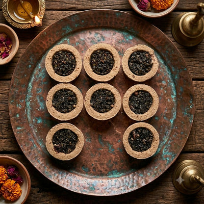 Nine round sambrani cups with dark contents arranged on a circular copper tray with floral accents.