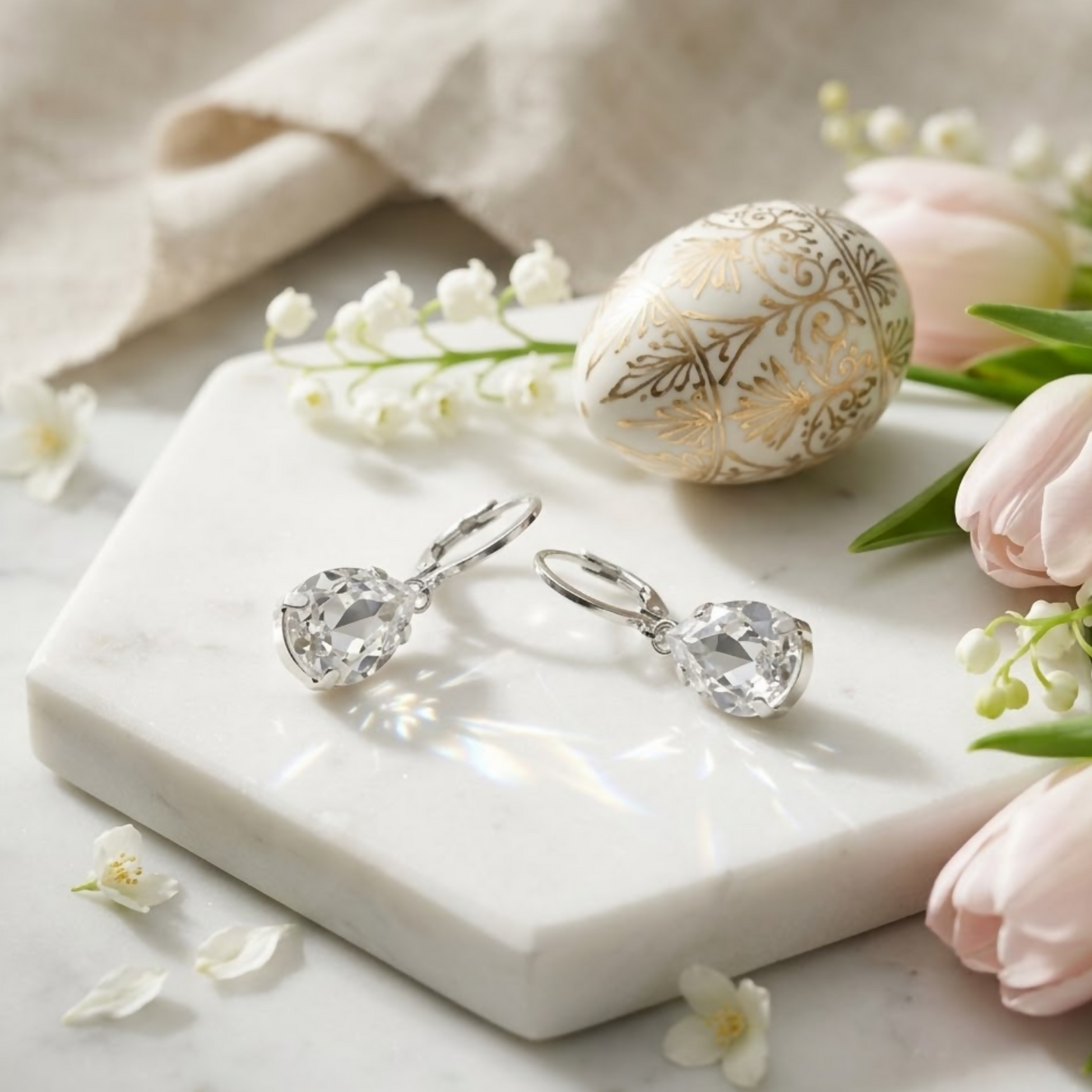Silver teardrop crystal earrings rest on a white marble slab next to a decorated egg and pink tulips.