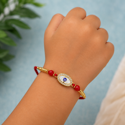 A child's hand wears a red thread bracelet with a golden oval evil eye charm, white stones, and red beads, against a blurry